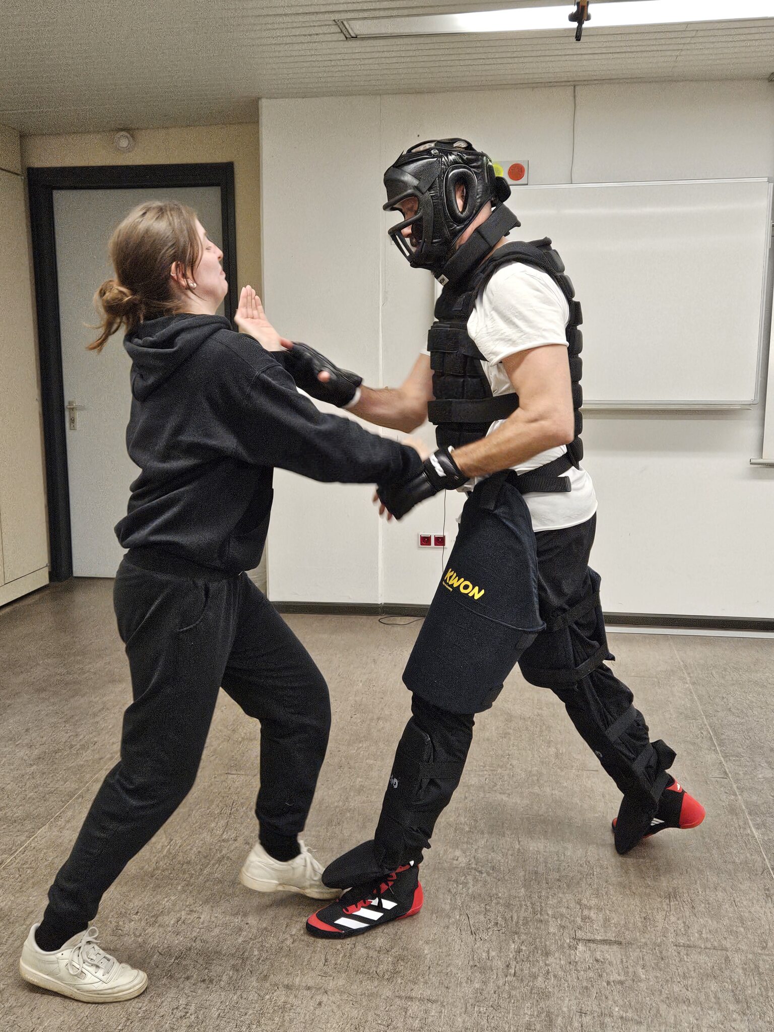Eine Person in Schutzausrüstung übt Selbstverteidigung mit einer anderen Person in einem Trainingsraum.