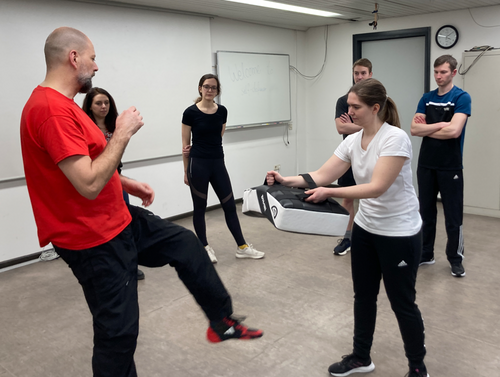 Trainer zeigt eine Technik im Kampfsporttraining mit mehreren Teilnehmern in einem Übungsraum.
