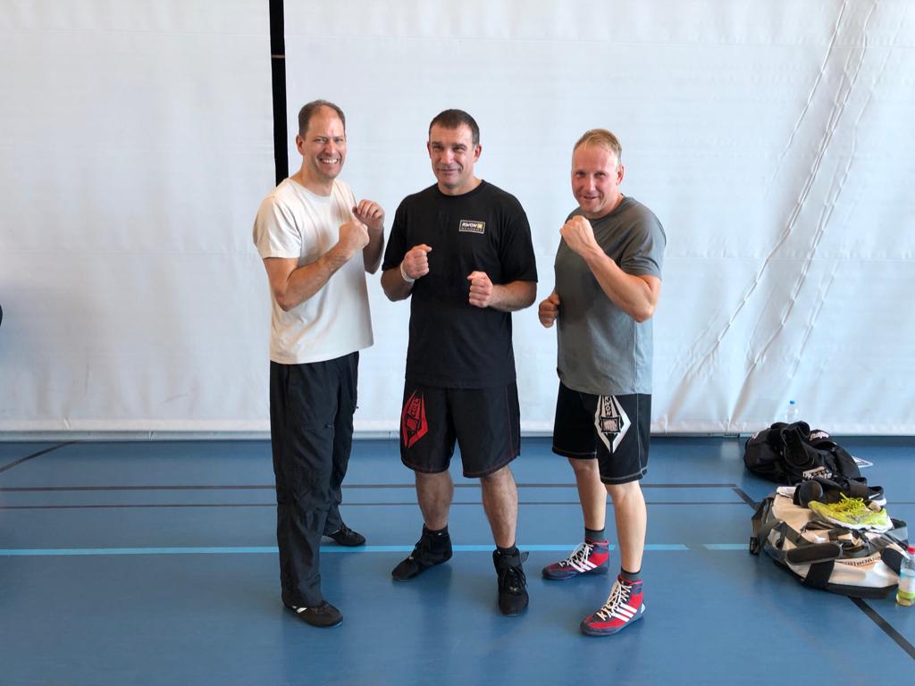 Drei Männer in Trainingskleidung stehen in einer Kampfsporthalle und posieren für ein Foto.