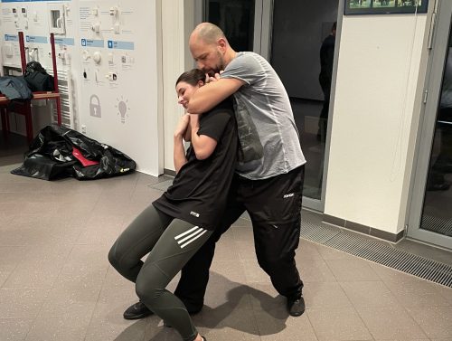 Zwei Personen üben Wing Chun Techniken in einem Trainingsraum, eine Person hält die andere in einer Umklammerung.